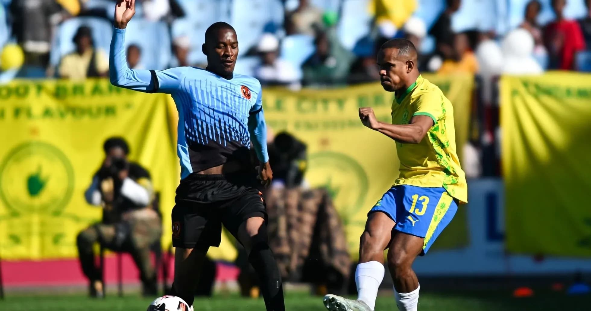 Player in yellow kicking ball, defender in blue nearby Player in yellow kicking ball, defender in blue nearby